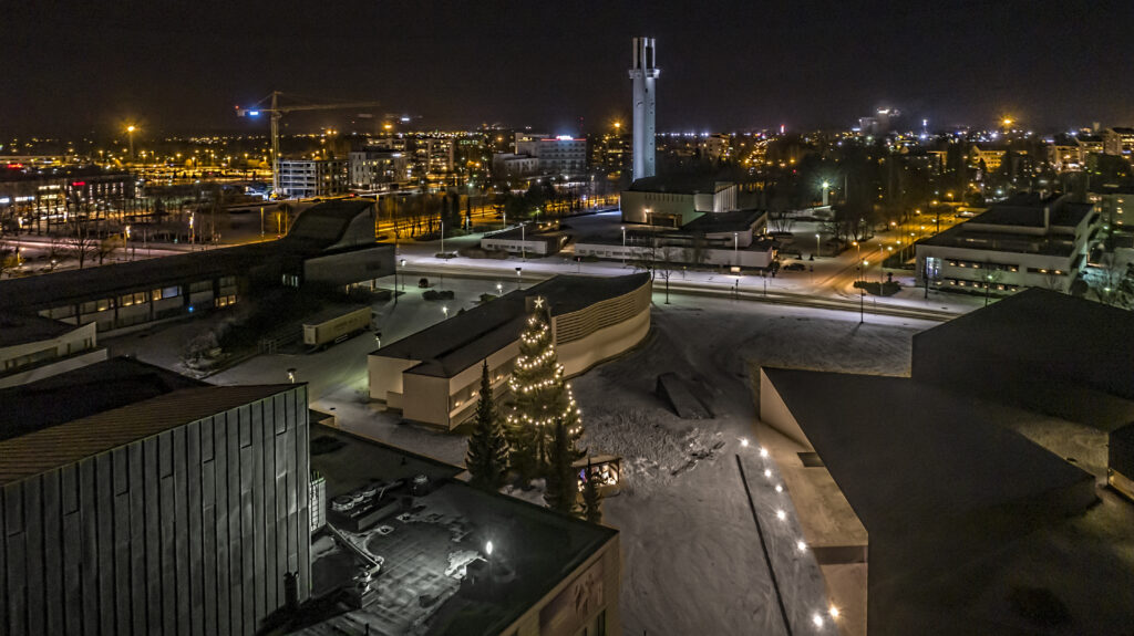 Pimeässä kaupunkikuvassa näkyy valoja ja joulukuusi sekä rakennuksia.